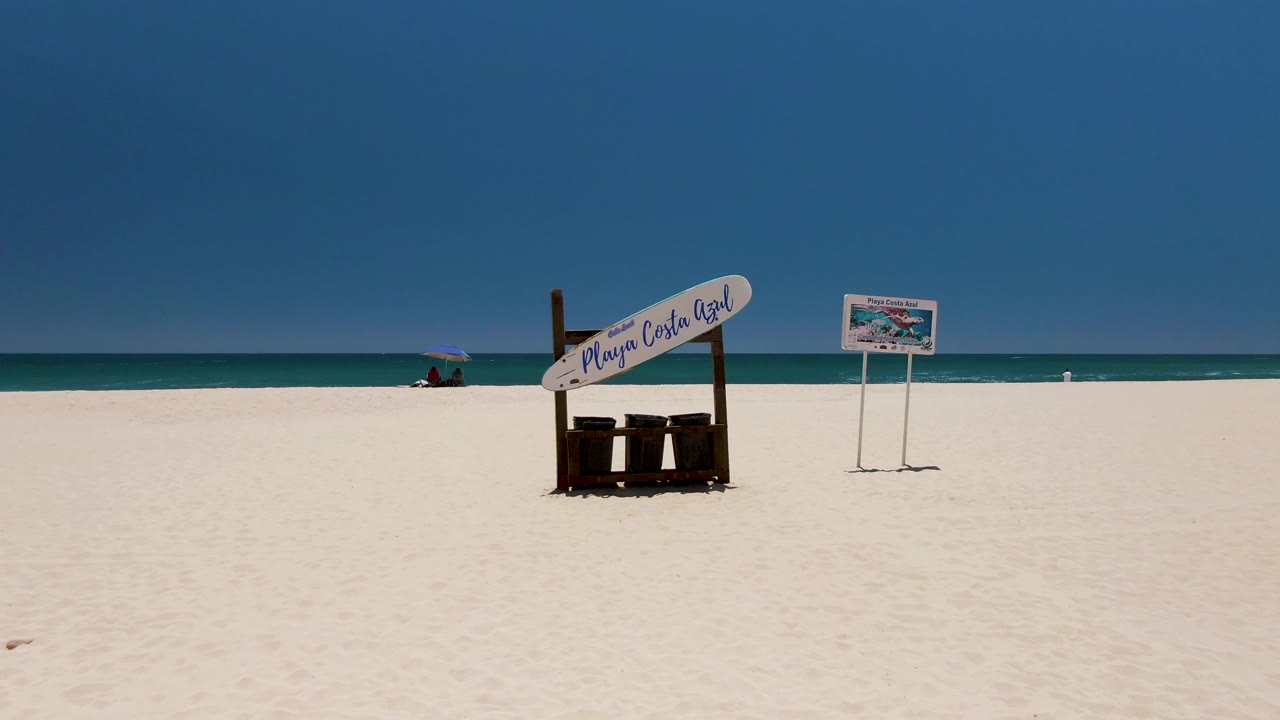 At Costa Azul Beach in Los Cabos, the wooden sign and separate posted sign stand on empty sand with the turquoise sea, low waves, and a distant umbrella along the horizon line.