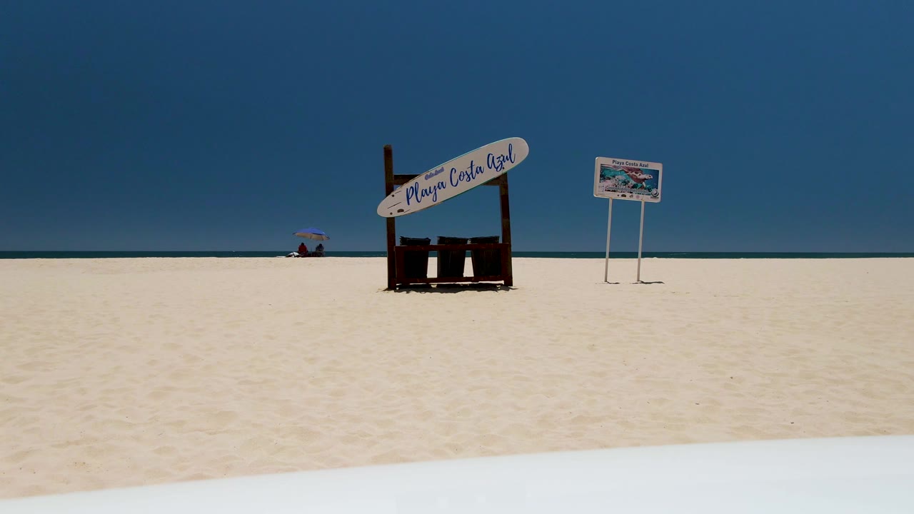 At 0:00, Costa Azul Beach in Los Cabos shows white sand, a Playa Costa Azul sign, a small beach board, blue ocean, clear sky, and a distant person carrying a red umbrella.