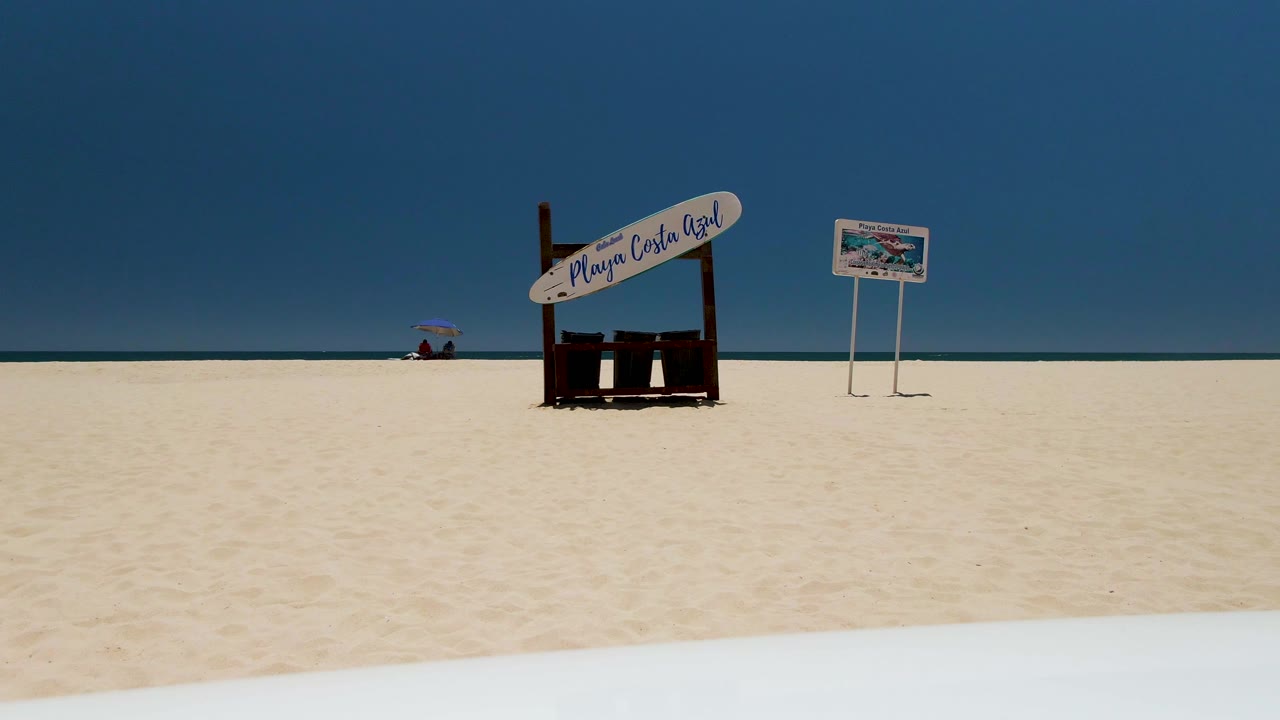 Playa Costa Azul sign and small board on white sand with a distant person under a red umbrella near the waterline.