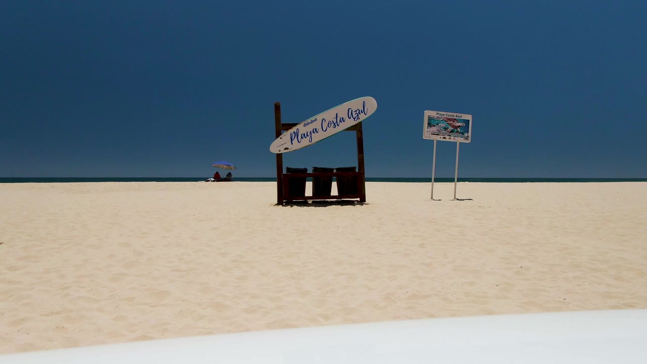At 0:02, Costa Azul Beach appears as a wide static-looking view with pale sand in front, the Playa Costa Azul sign and small board midframe, and the umbrella walker still distant by the water.
