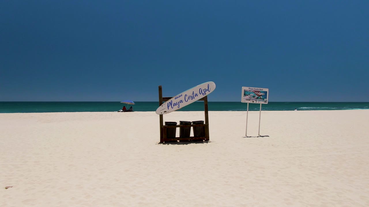 Wide view of Costa Azul Beach in Los Cabos with white sand, a large beach sign, a smaller sign, calm ocean, clear blue sky, and a tiny distant beachgoer near the water.
