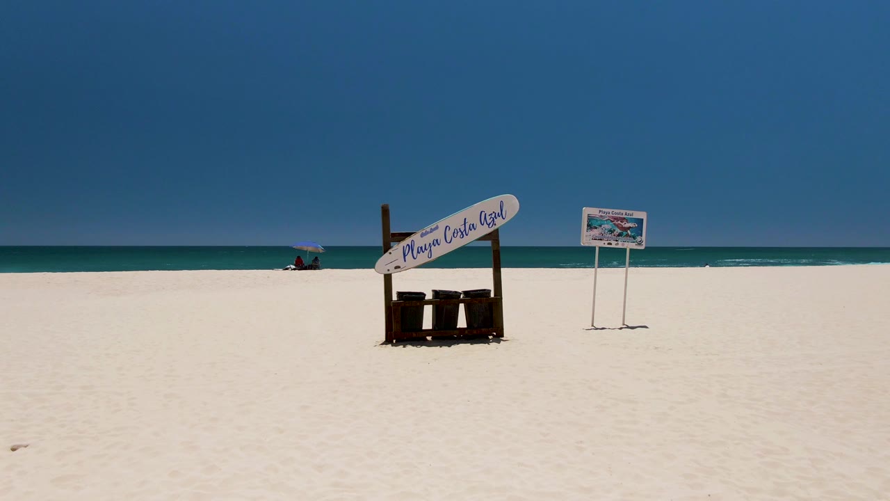 Costa Azul Beach sign sits slightly left of center on white sand, with a smaller sign to the right, turquoise ocean behind, and a tiny distant beachgoer near the shoreline.