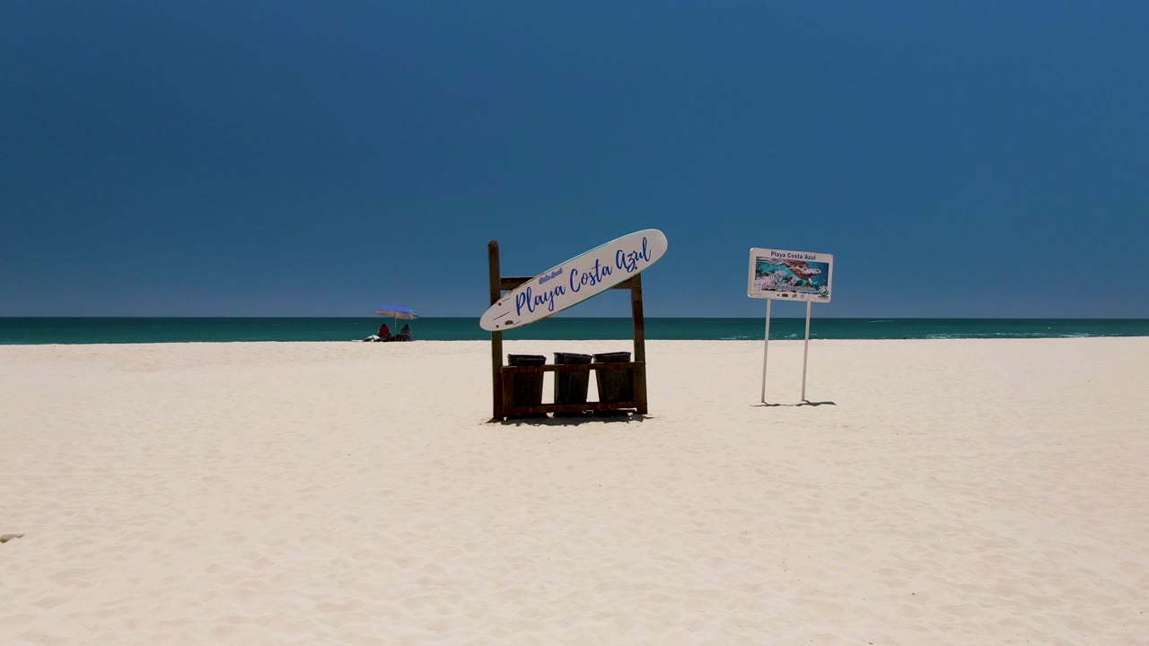 A wide Costa Azul Beach frame shows the large sign and smaller sign spaced across the sand, with calm ocean bands and a tiny distant umbrella near the waterline.