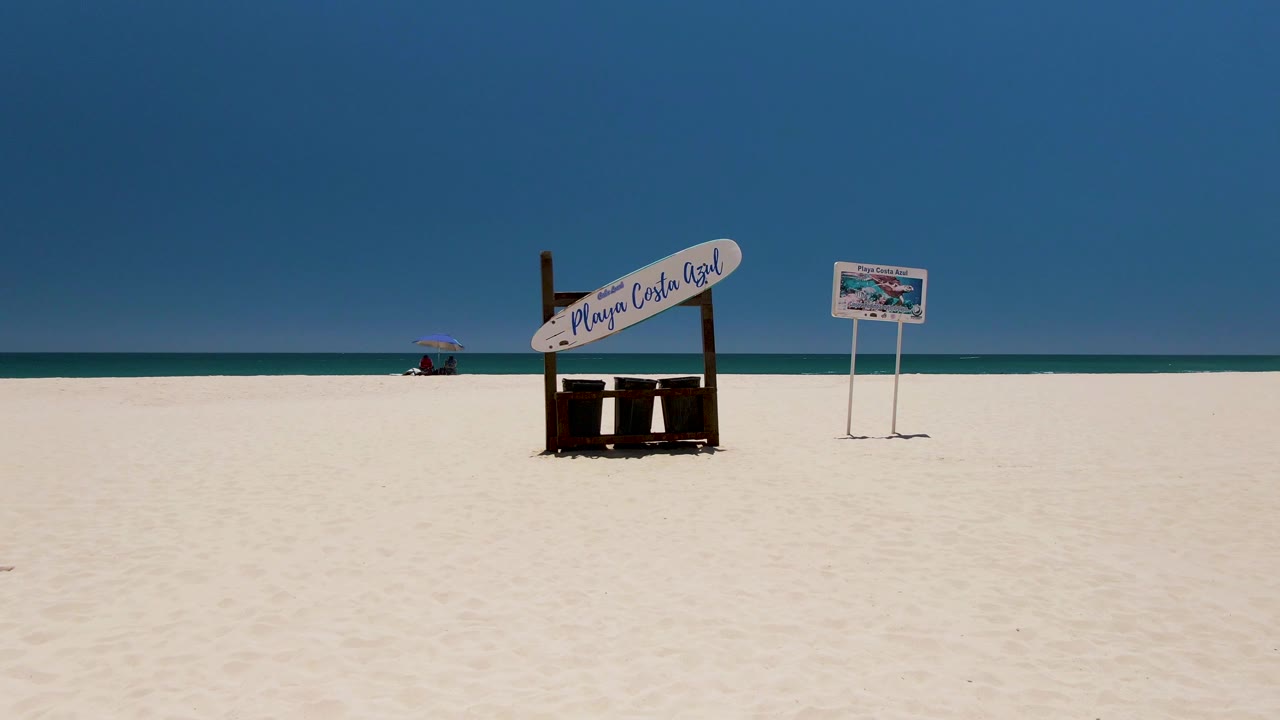 Large beach sign and smaller sign on bright sand beside calm sea.