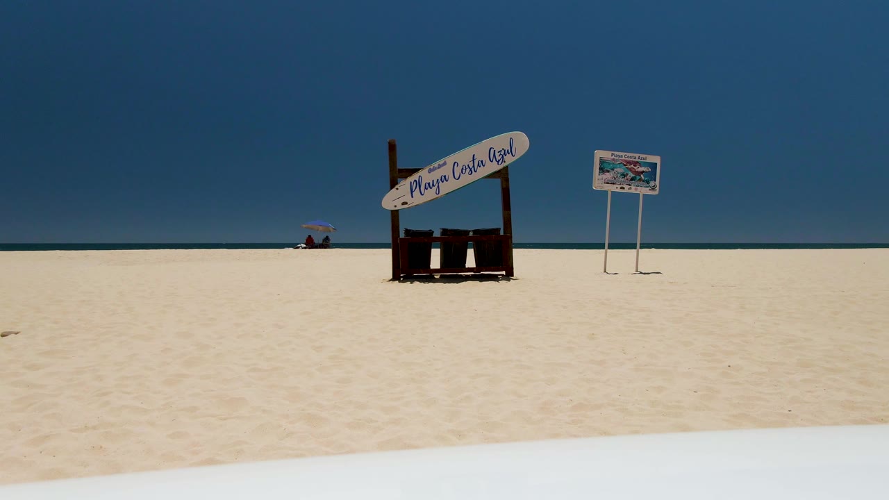 Opening frame at Costa Azul Beach in Los Cabos showing pale sand, a wooden beach sign, a small signboard, calm ocean, clear blue sky, and no visible people.