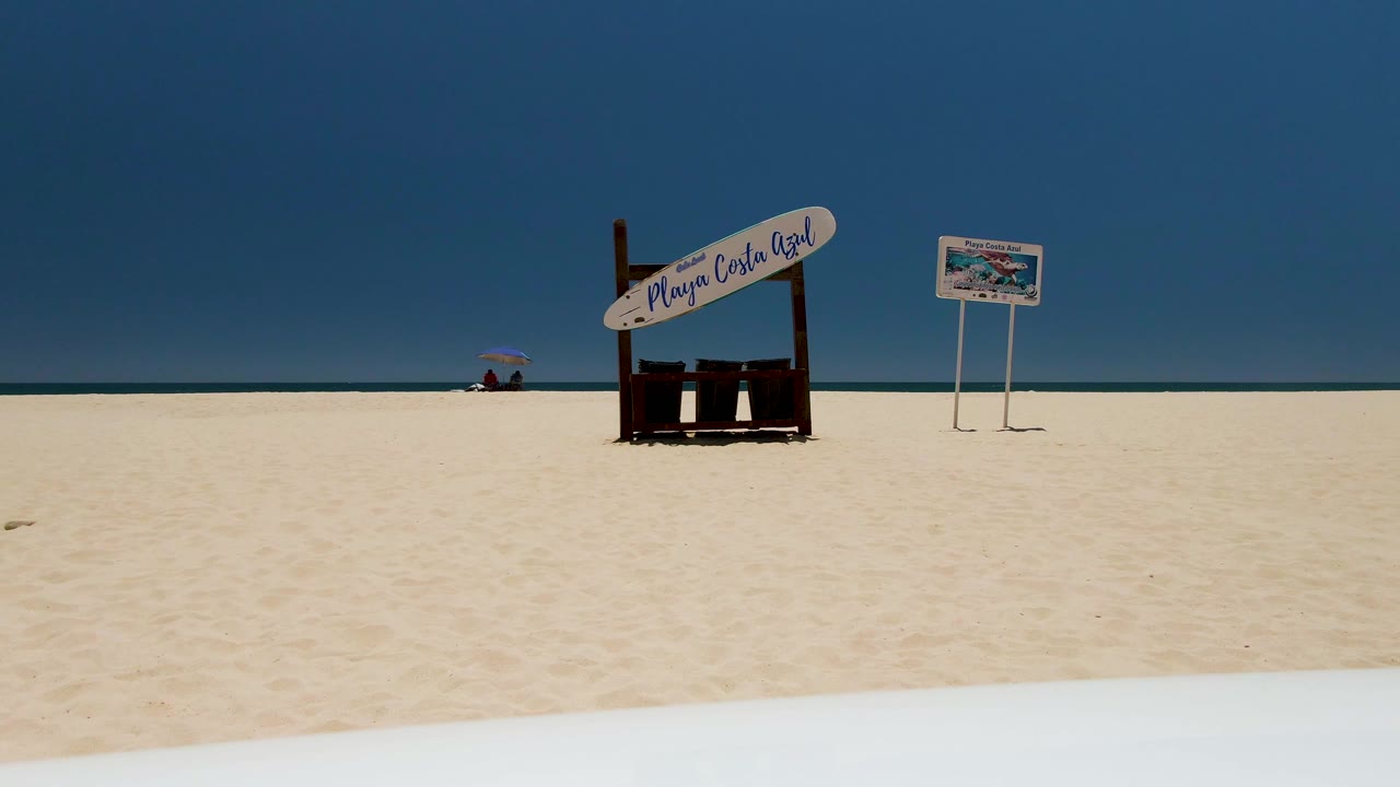 One second into the clip, the static Costa Azul Beach view holds on the wooden sign and smaller signboard against the ocean horizon, with no visible people.