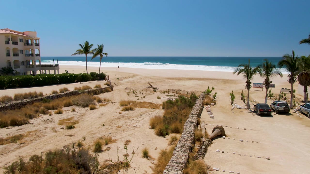 Aerial frame from San Jose del Cabo showing tan dunes, a sandy road, palm trees, a beachside building at left, white surf, blue ocean, and no visible people.