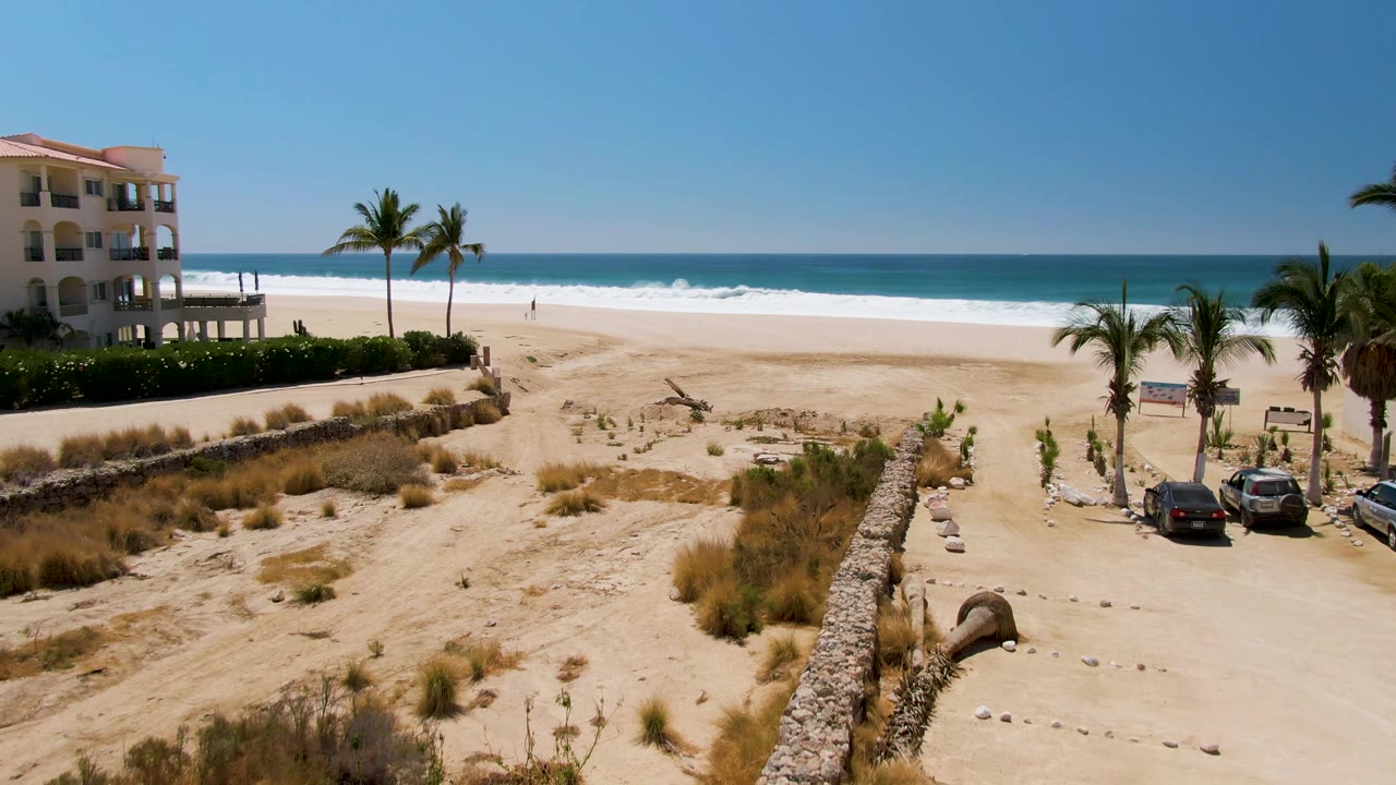 San Jose del Cabo aerial view with the beachside building near center-left, parked cars beside palms, dune plants below, surf bands across the beach, and no visible people.