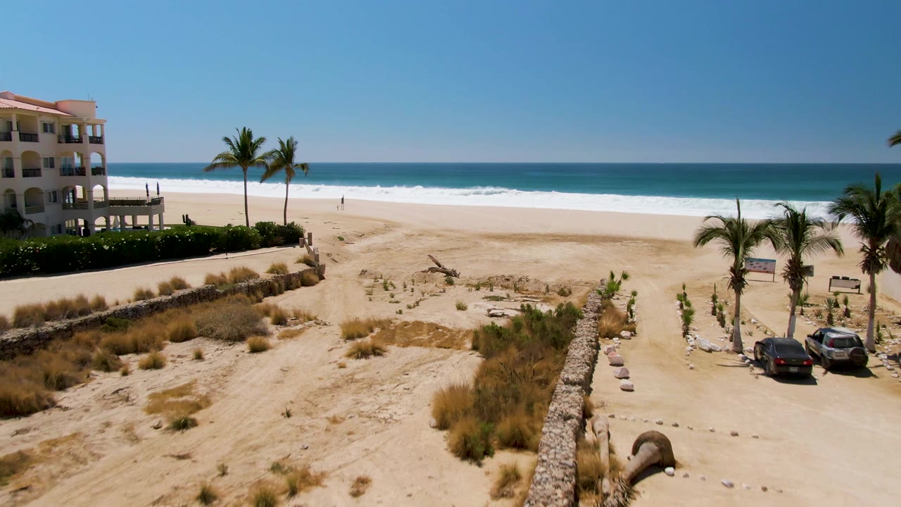 San Jose del Cabo drone frame showing the building shifted toward the left, palms and parked cars on the right, planted dunes in front, ocean surf behind, and no visible people.
