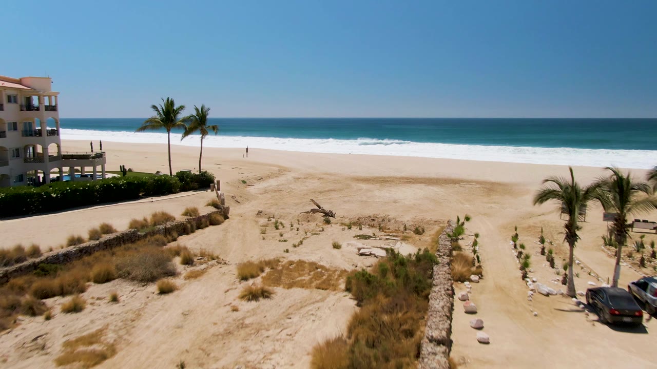 Beach dunes and palms beside a building with ocean waves in the background.