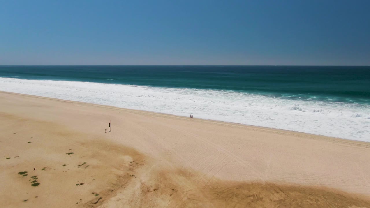 San Jose Del Cabo aerial shoreline frame with the palm trees mostly gone, open tan sand below, white waves across the middle, blue water beyond, and distant people near the beach.