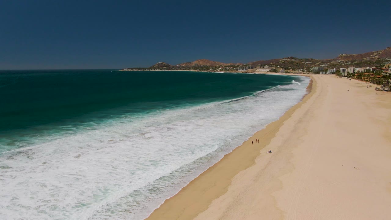 Aerial view in San Jose del Cabo showing dark teal ocean on the left, white foam reaching a broad sandy beach, coastal buildings, hills, and tiny beachgoers in the distance.