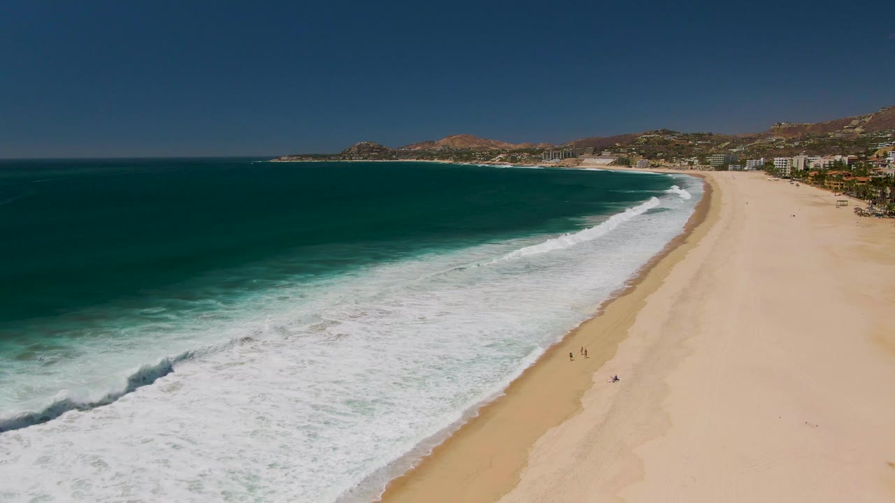 Aerial view over San Jose del Cabo coastline as a darker wave line breaks into white foam near the lower left, with the sandy beach, small beachgoers, buildings, and hills beyond.