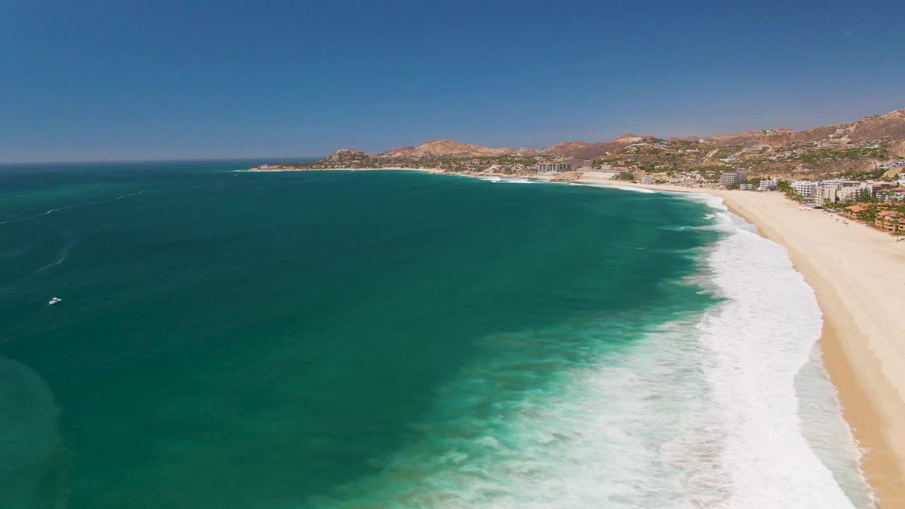 Aerial view of turquoise ocean waves beside a sandy beach and distant coastal hills.