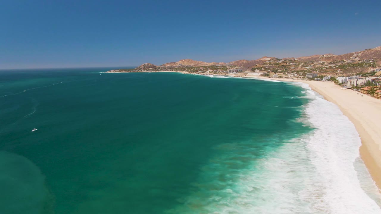 Aerial view over San Jose Del Cabo with more open turquoise water in the center, the foamy shore break running down the right side, coastal buildings on the distant sand, hills beyond, and no visible people.
