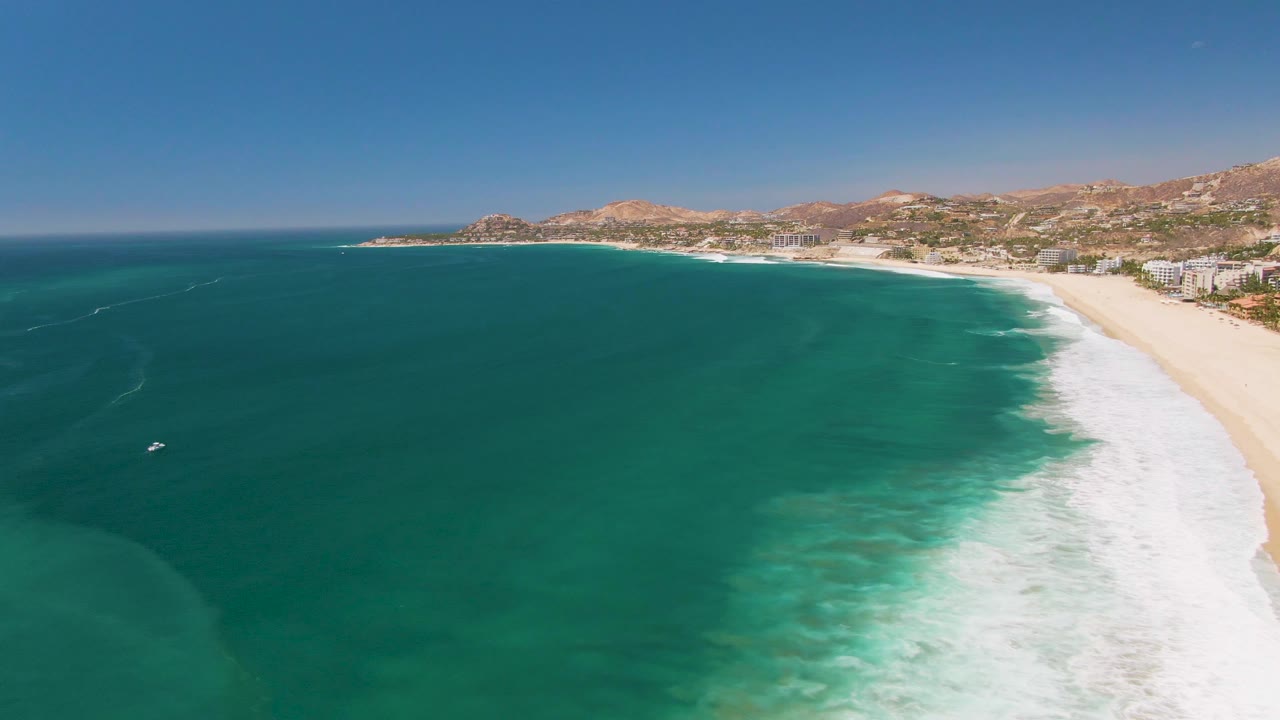 San Jose Del Cabo aerial coastline frame with the beach compressed along the right edge, darker turquoise water across the center, a white surf band, distant buildings, tan hills, and no visible people.