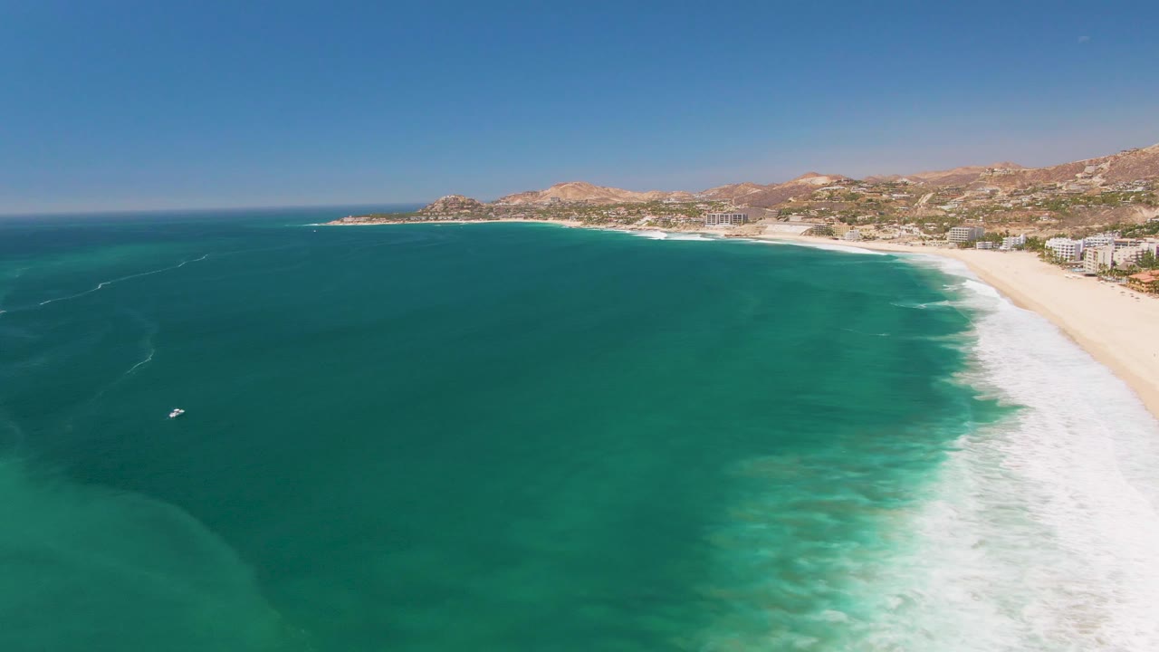 Aerial view of the San Jose Del Cabo coast with a small white boat more apparent on the left water, pale underwater color near the lower edge, white surf at right, distant buildings, hills, and no visible people.