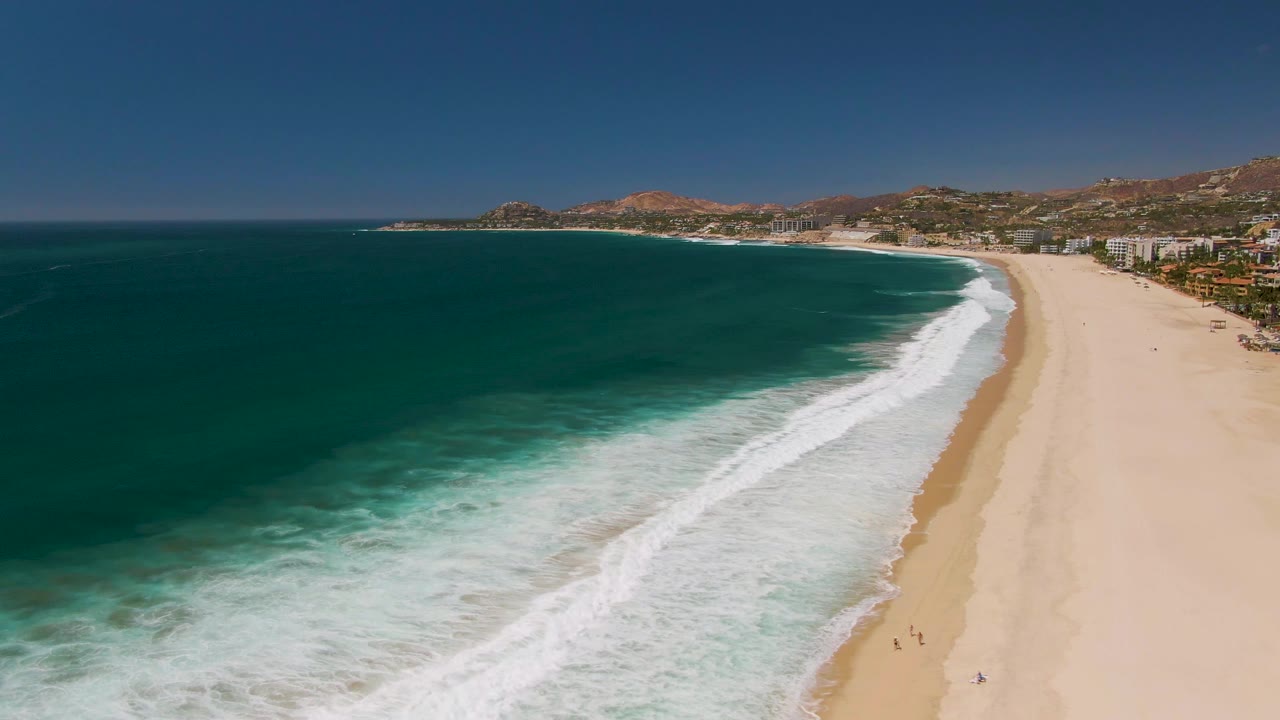 Aerial frame from San Jose Del Cabo showing deep blue water, white surf, a wide sandy beach, coastal hills, beachfront buildings, and tiny distant beachgoers on the sand.