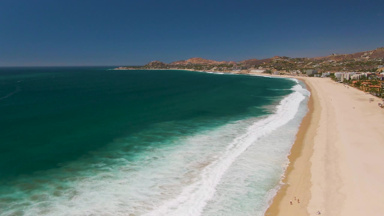 Drone view of white surf curling along a broad sandy shoreline.