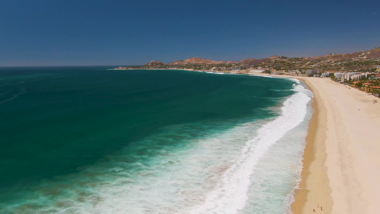 San Jose Del Cabo aerial frame showing turquoise shallows, a broad white surf band, empty-looking sand in the foreground area, and developed coastline beneath dry hills in the distance.