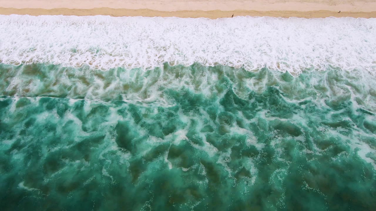 Top-down view of green ocean water and white foam breaking onto sand in San Jose del Cabo, with no visible people.