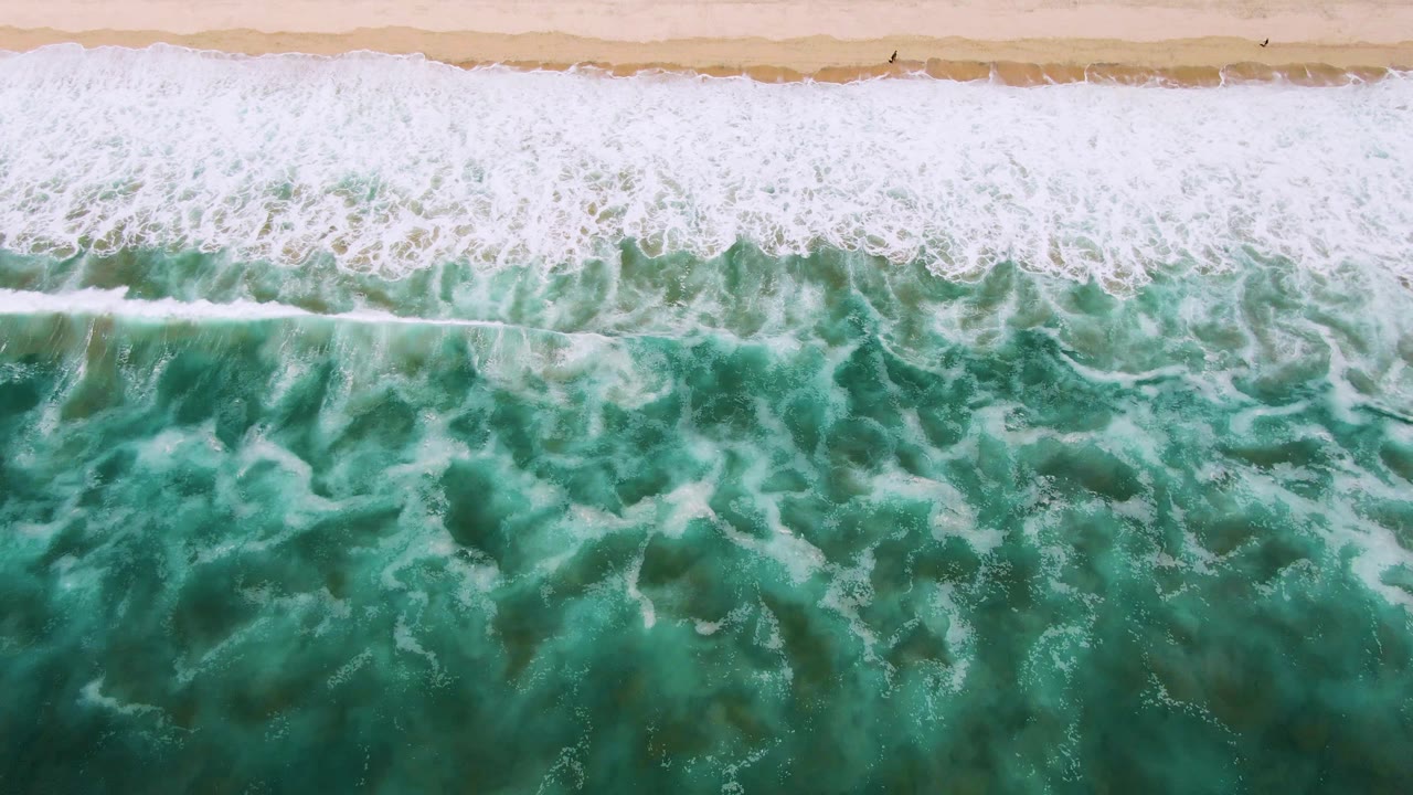 Top-down San Jose del Cabo beach view with a dense white breaker across the sand edge and green water below, with no visible people.