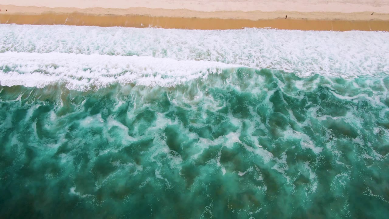 Overhead view in San Jose Del Cabo, Mexico, with turquoise water and white foam approaching a narrow sandy beach at the start of the clip, no visible people.