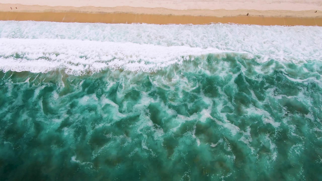 Overhead San Jose Del Cabo shoreline frame with a thicker white foam band spreading across turquoise water below the sandy beach, no visible people.