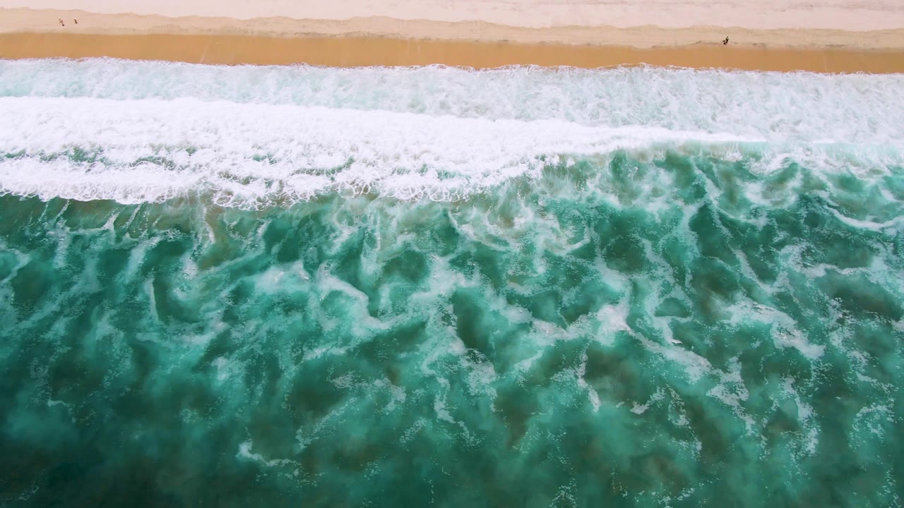 Aerial view of the San Jose Del Cabo coast with textured turquoise water, scattered white foam, and tan sand running across the top of the frame, no visible people.