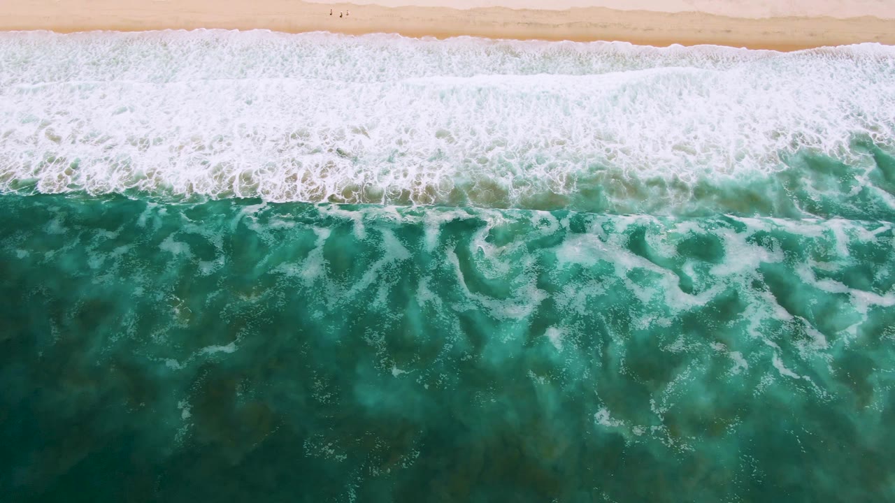 Top-down view of turquoise surf and white foam approaching pale sand in San Jose Del Cabo, Mexico, with tiny distant beach figures near the upper edge.
