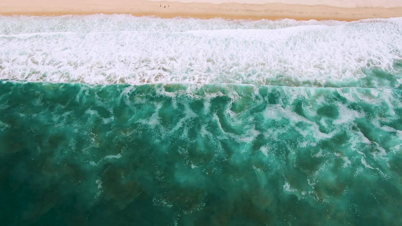 Whitewater spreads across turquoise ocean beside a sandy shoreline.