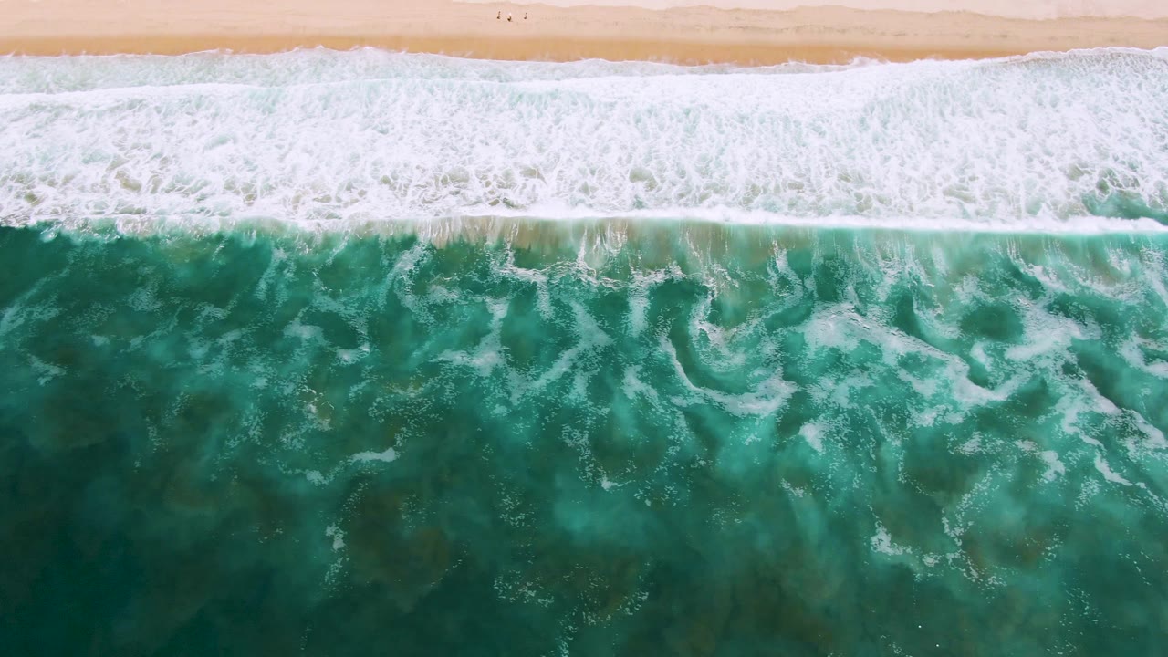 Top-down San Jose Del Cabo surf view with dense white foam near the sand and textured turquoise water filling the lower frame, with no visible people in this frame.