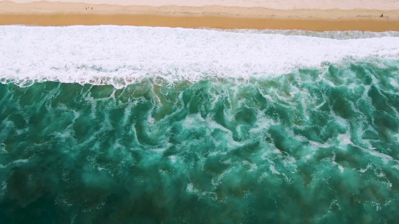 Aerial San Jose Del Cabo shoreline frame with white surf spreading wider across textured green water below a narrow strip of pale beach, with no visible people.