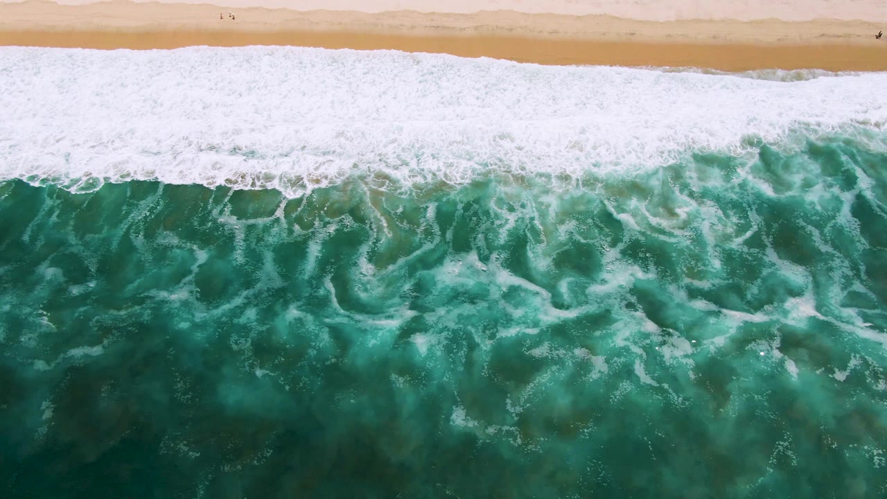 Top-down frame in San Jose Del Cabo, Mexico, with a long foamy wave edge running parallel to the sandy beach and darker green water below, with no visible people.