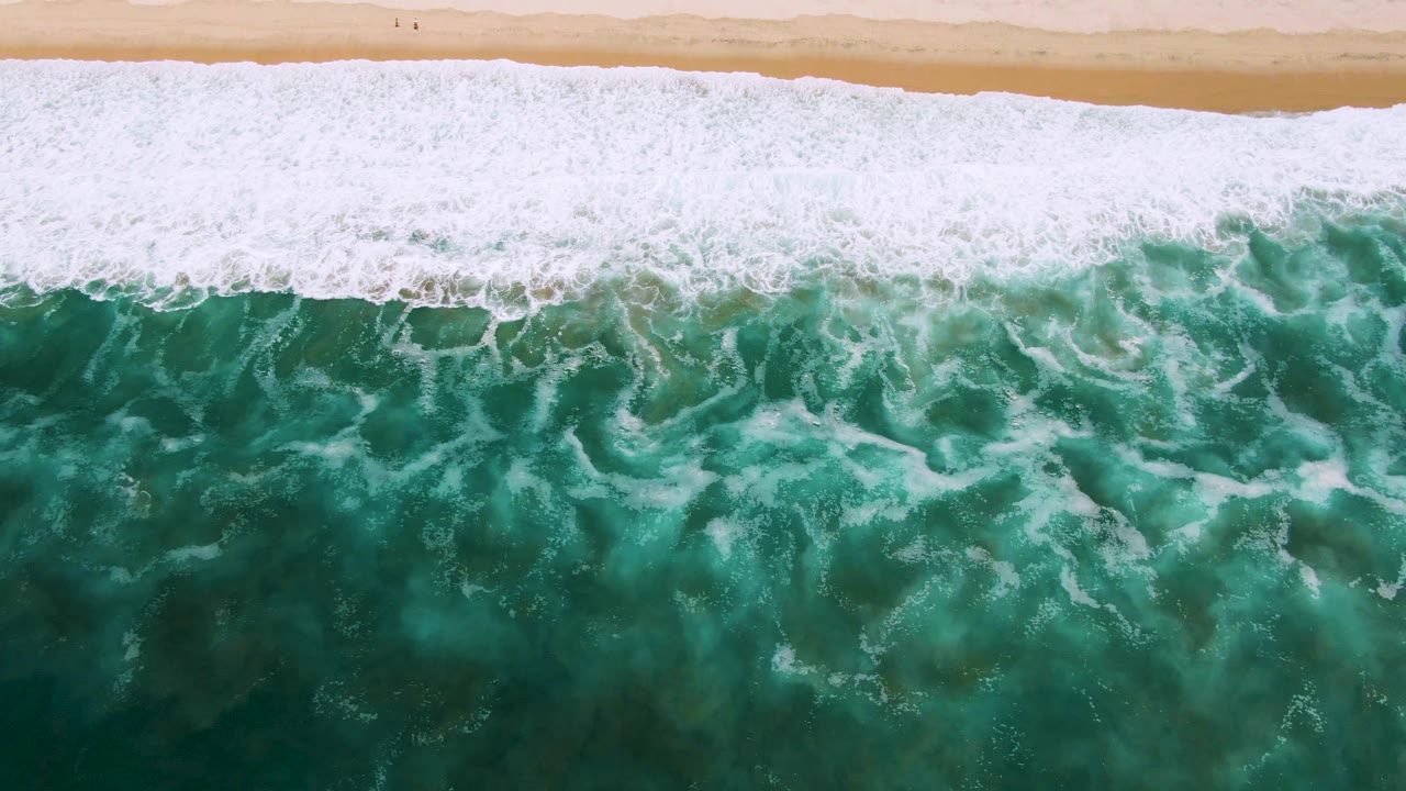 Overhead San Jose Del Cabo frame showing dense white foam washing toward tan sand while mottled green ocean texture fills the lower portion, with no visible people.