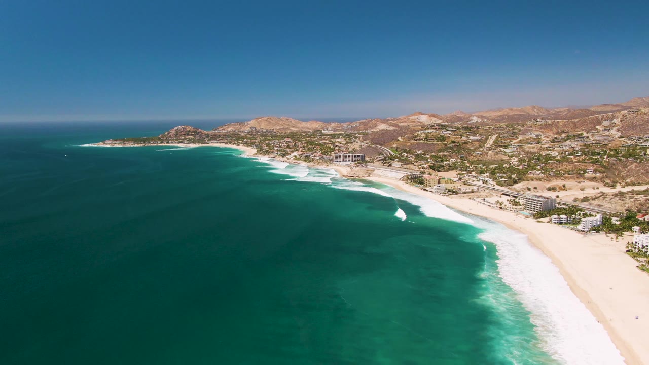 Aerial view of San Jose del Cabo coastline with turquoise water in the foreground, white surf on a sandy beach, coastal buildings, dry hills, clear blue sky, and no visible people.