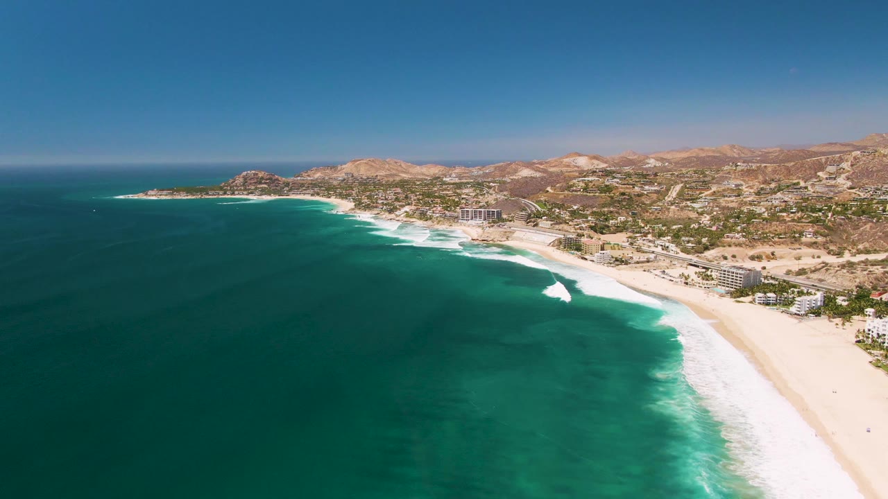 Aerial San Jose del Cabo shoreline with deep turquoise water taking most of the frame, white foam bands near the sand, buildings along the coast, arid hills inland, and no visible people.