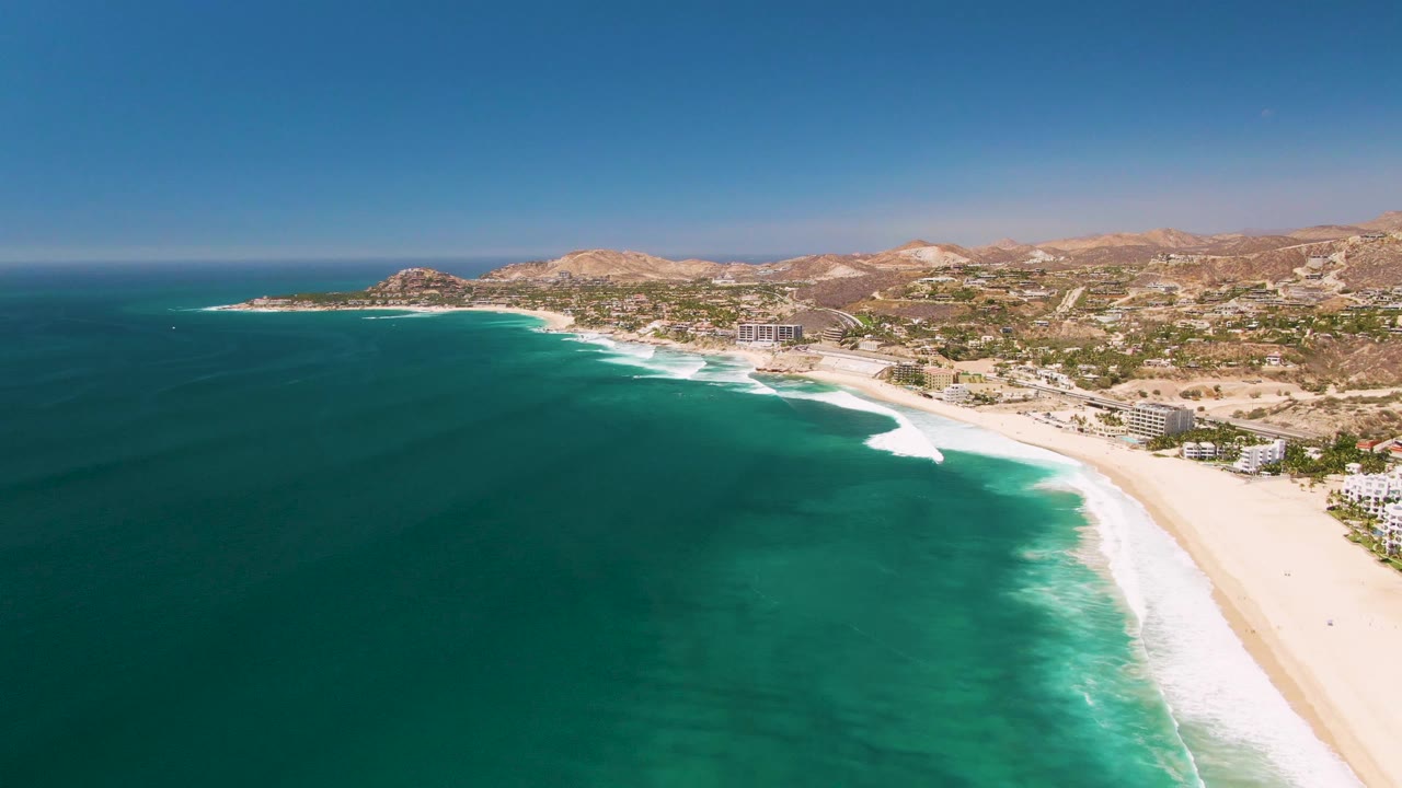 Aerial view of San Jose Del Cabo coastline with turquoise water in the foreground, white surf along the beach, hillside buildings in the distance, and no visible people.