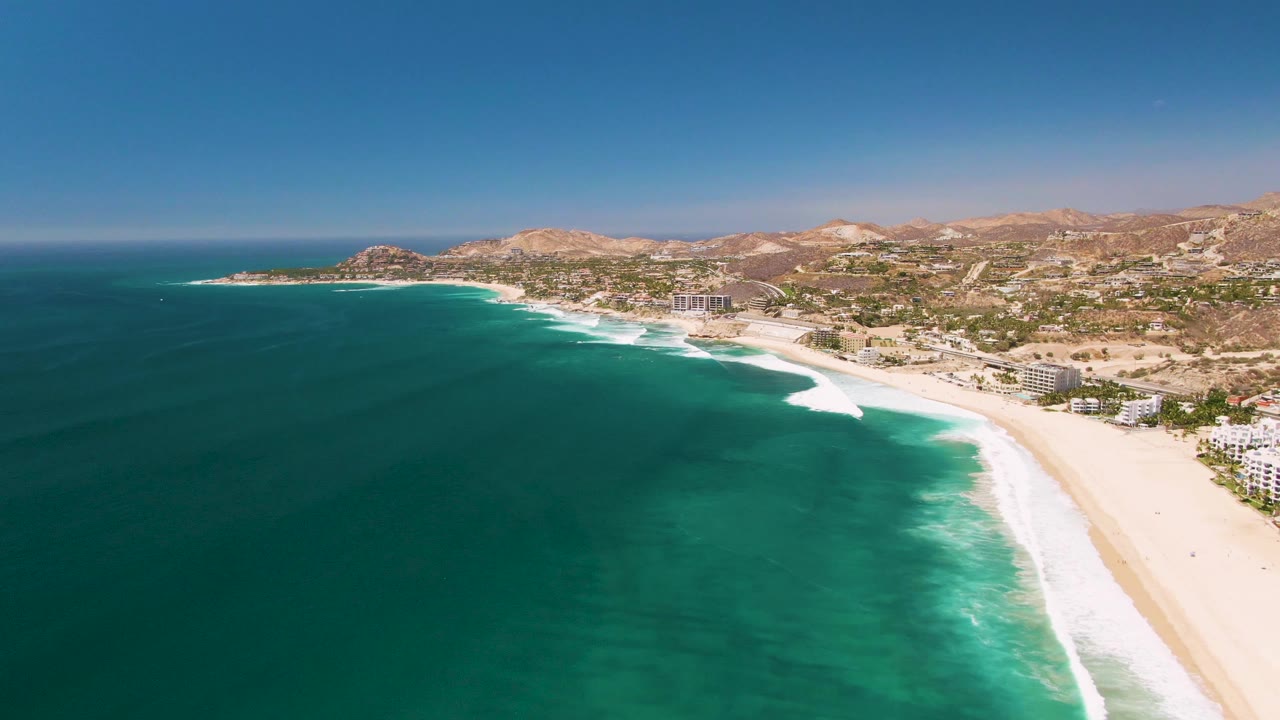 Wide aerial beach view with breaking waves and hillside development beyond.