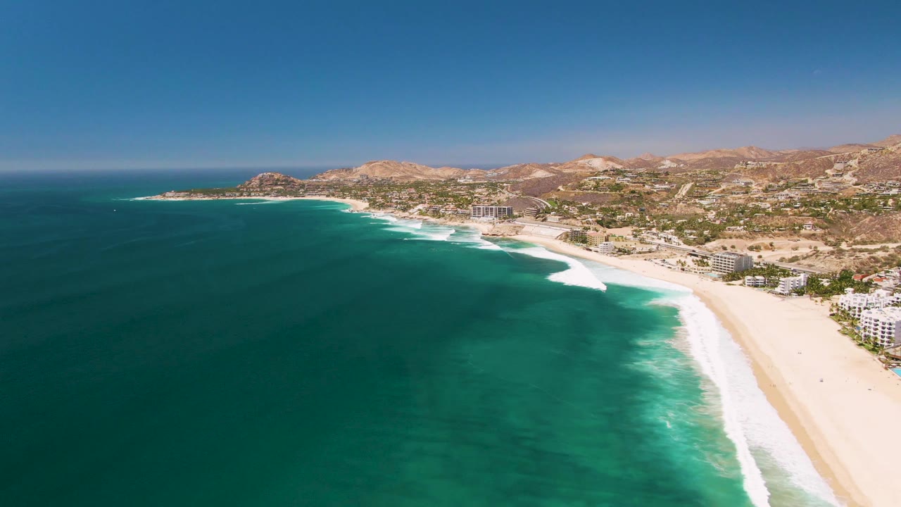 Drone-style view along the San Jose Del Cabo shoreline, with broad turquoise water, a bright surf band, beachfront buildings near the lower right edge, and no visible people.