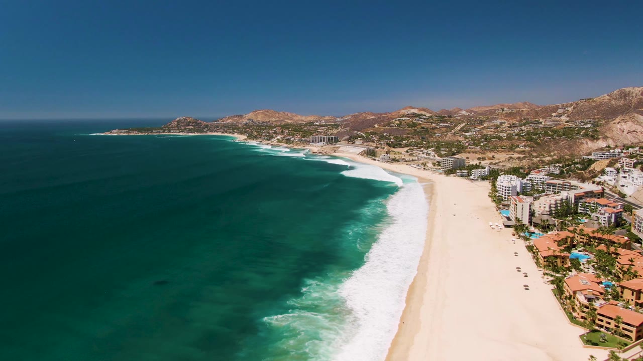 Aerial frame over San Jose Del Cabo showing turquoise ocean on the left, a pale sandy beach, beachfront buildings to the right, dry hills in the distance, and no visible people.