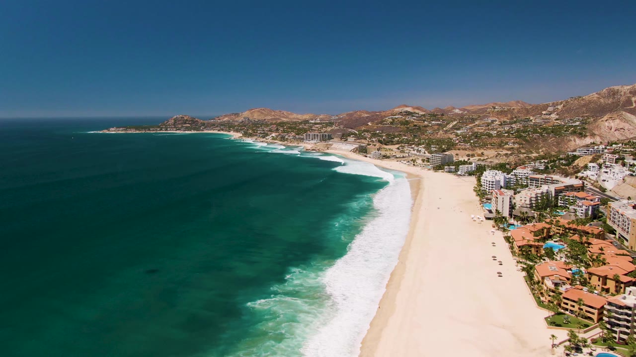 Wide drone view of waves rolling toward a sandy beach beside coastal buildings.