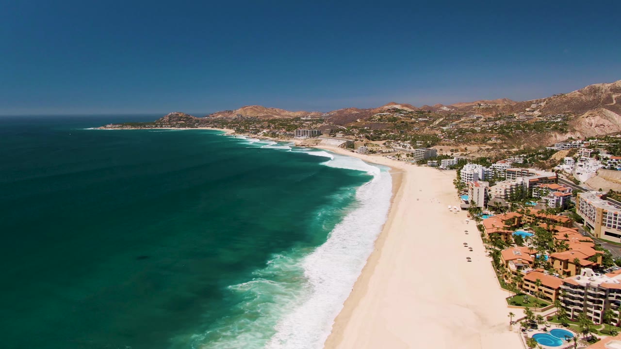 Aerial frame in San Jose Del Cabo with the coastline curving into the distance, surf breaking along the sand, resort-style buildings at frame right, hills beyond, and no visible people.