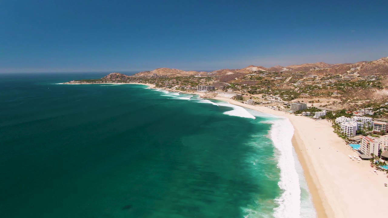 Aerial view of turquoise surf rolling onto a sandy beach beside coastal buildings and hills.