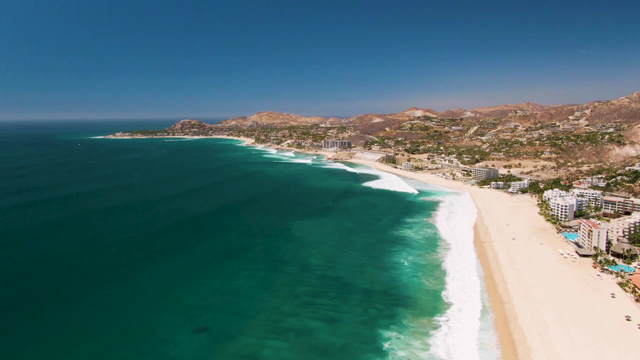 One second into the San Jose del Cabo clip, the aerial view holds on a long sandy shoreline with white foam bands, blue-green water, beachfront buildings, distant hills, and no visible people.