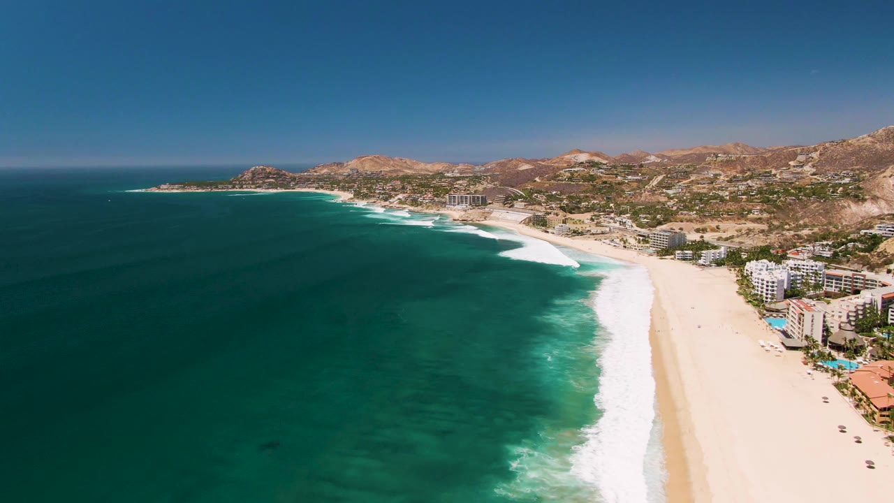 At 0:02, the San Jose del Cabo aerial frame shows a broader stretch of curling white surf, open blue water, pale sand, clustered coastal buildings on the right, arid hills, and no visible people.