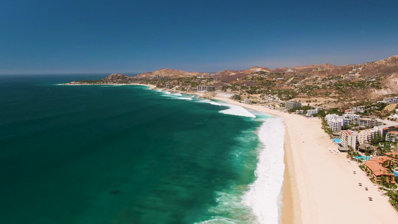 At 0:03, the aerial San Jose del Cabo view places more deep blue water in the foreground, with foamy surf wrapping the beach, coastal buildings along the right edge, hills beyond, and no visible people.
