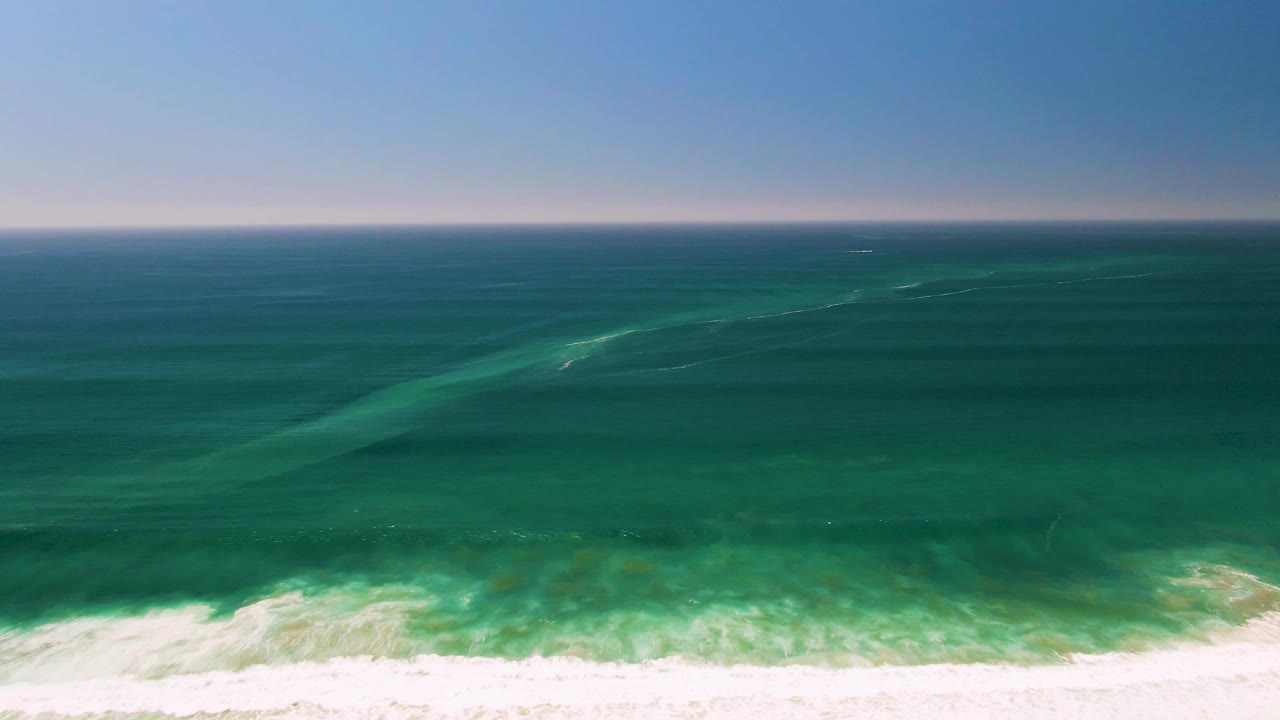 San Jose Del Cabo shoreline frame with a level ocean horizon, green-blue water bands, and white foam spreading across the lower edge, no visible people.