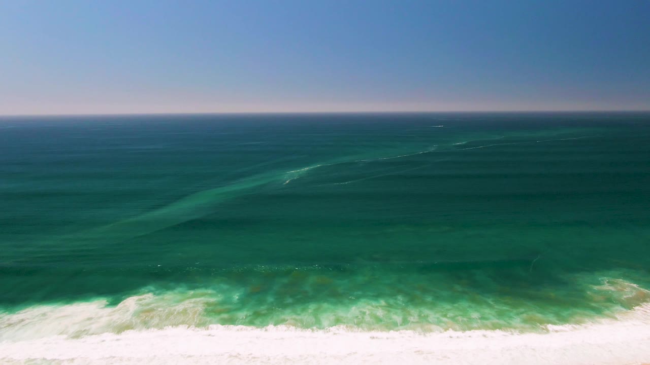 Aerial coastal view from San Jose Del Cabo, Mexico, where darker offshore water fades into turquoise shallows and white foam curls over the sand, no visible people.