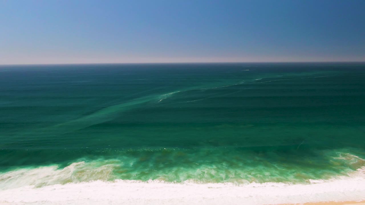 Wide aerial frame of the San Jose Del Cabo coast with broad green ocean surface, a thin clear horizon, and white surf stretched along the lower sand, no visible people.